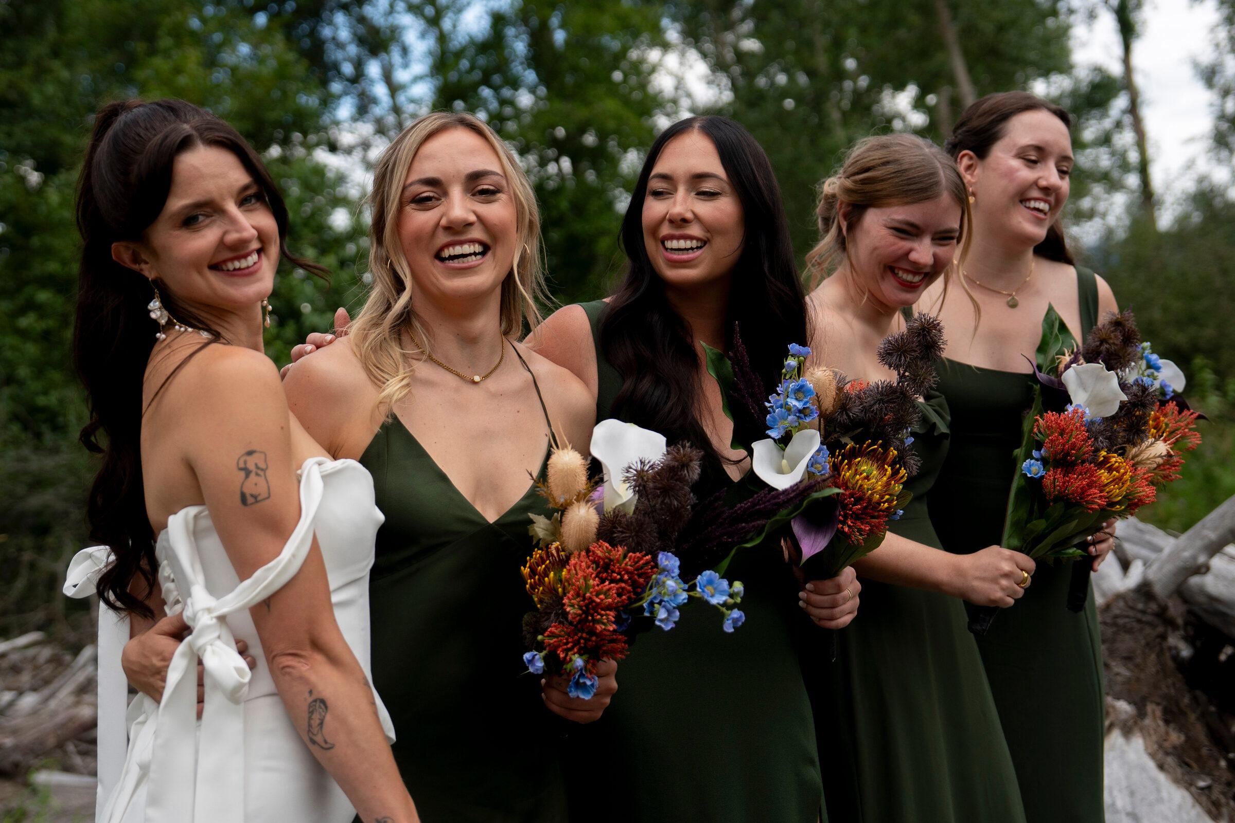 Bride and four bridesmaids in forest green laughing by the river, protea and calla lily bouquets in hand