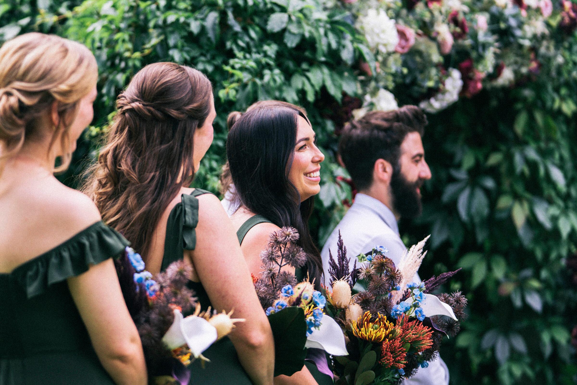 Bridal party in profile beside the greenery-and-blossom ceremony wall